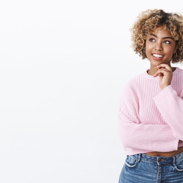 Attractive young african-american female student with blond hair holding hand on chin and looking delighted at upper left corner as thinking, posing thoughtful pleased with idea came to mind Femme qui réfléchit