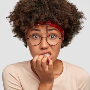 Photo of embarrassed dark skinned woman looks anxiously at camera, bites finger nails, has puzzled nervous facial expression, curly dark hair, poses against white background. Emotions concept jeune fille qui se ronge les ongles